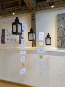 3 black lanterns hanging in gallery with white paper - there are words on the windows of the lanterns and on the papers